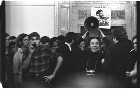 A young Mark Rudd speaking into a microphone at a rally with a portrait of Che Guevara in the background.