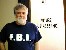 Mark Rudd wearing an F.B.I. t-shirt and standing in front of a door with the words 'Future Business Inc' printed on it.