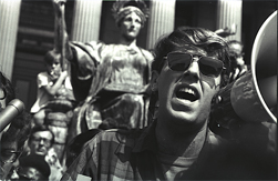 A young Mark Rudd wearing sunglasses and yelling into a megaphone.